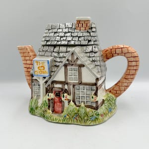 Peut inclure: Théière en céramique en forme de cottage, avec un toit en tuiles grises et une cheminée en briques. La théière a une anse et un bec en briques, et une enseigne indiquant "The Crown". La base est ornée de feuillage vert.