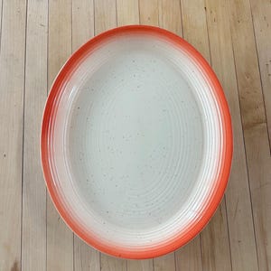 May include: Oval ceramic serving platter with a white center and a gradient orange rim. The platter has a speckled surface and concentric ring design. A decorative piece for serving food.