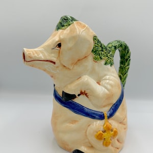 May include: A ceramic pig-shaped pitcher with a cream-colored body, adorned with green leaf accents and a blue ribbon. The pitcher features a gold cross pendant and a green handle, perfect for serving beverages.
