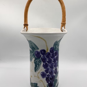 May include: A white ceramic vase with a woven handle. The vase features a hand-painted design of purple grapes and green leaves. The handle is made of light brown woven material.