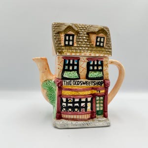 Peut inclure: Théière en céramique en forme de boutique, avec un toit brun et deux petites fenêtres. La façade de la boutique est beige avec des détails rouges et verts, et le texte "THE OLDSWEETSHOP" est affiché au-dessus des fenêtres. Le bec et la anse sont de couleur pêche clair.
