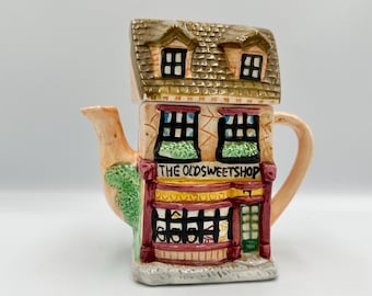 May include: A ceramic teapot shaped like a quaint shop, featuring a brown roof with two small windows. The shop's facade is beige with red and green accents, and the text "THE OLDSWEETSHOP" is displayed above the windows. The spout and handle are a light peach color.