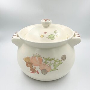 May include: A cream-colored ceramic pot with a matching lid, featuring two side handles and a knob. The exterior is decorated with watercolor-style illustrations of various vegetables, including tomatoes, peas, and mushrooms, with small floral accents.
