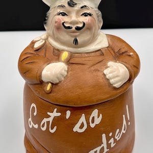 May include: A ceramic cookie jar shaped like a smiling chef, wearing a white hat and brown apron. The jar is brown with the words "Lot's a goodies!" written in white script.