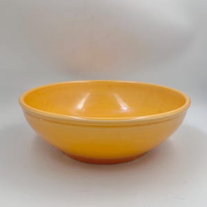 Puede incluir: Un cuenco de cerámica amarillo brillante con forma redondeada. El cuenco tiene un acabado liso y brillante y una base ligeramente más oscura de color naranja. El cuenco está vacío y descansa sobre una superficie blanca.