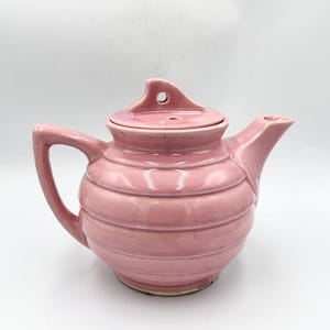 May include: A vintage pink ceramic teapot with a lid. The teapot has a handle and a spout. The body of the teapot is decorated with horizontal ridges. The lid has a small handle with a hole. The teapot is on a white background.