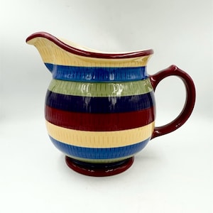 May include: A ceramic pitcher featuring a rounded body and a curved handle, decorated with horizontal stripes in cream, blue, sage green, and deep red. The surface has a subtle vertical ribbed texture throughout, and the base is a solid deep red.