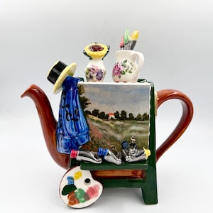 May include: A ceramic teapot shaped like an artist's easel, featuring a landscape painting on the front, a blue smock with a straw hat, a sunflower vase, paint brushes, tubes of paint, and a colorful palette attached to the dark green frame.