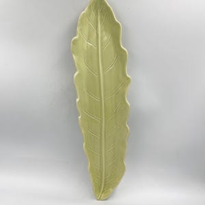 May include: A light green, leaf-shaped ceramic serving dish. The dish has a long, slender shape with a textured surface and detailed veining. The edges are gently scalloped. The dish is approximately 15 inches long.