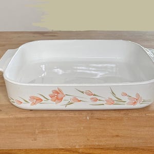 May include: White rectangular baking dish with handles, decorated with a floral pattern of peach-colored flowers and green leaves. The dish is made of ceramic and sits on a wooden surface.
