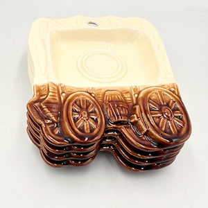 May include: A stack of six ceramic wagon-shaped plates. The plates are cream-colored with brown wagon wheel and barrel detailing. The top plate has a recessed center and a small hole for hanging.