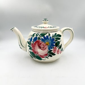 May include: A cream-colored ceramic teapot featuring a hand-painted floral design with a large pink rose, a bright blue daisy, and green leaves. The teapot includes a matching lid with a small knob, a curved spout, and a sturdy handle.