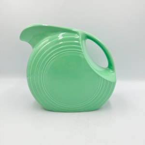 May include: A mint green ceramic pitcher with a rounded body and a curved handle. The pitcher has a spout for pouring and a series of concentric rings that decorate the exterior. The pitcher is a classic piece of kitchenware.