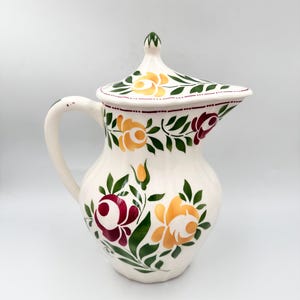 May include: Cream ceramic jug with lid, decorated with hand-painted floral patterns in yellow, burgundy, and green. The jug has a curved handle and a spout. The lid is topped with a green finial.
