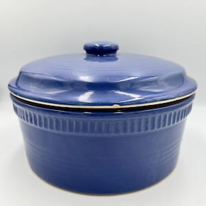 Puede incluir: Una fuente para horno de cerámica azul cobalto con tapa a juego. El plato tiene forma redonda con un exterior acanalado y un pequeño pomo en la tapa. El plato está diseñado para cocinar y servir.