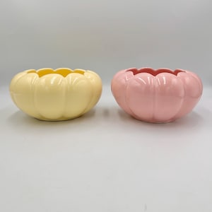 May include: Two ceramic bowls in a pumpkin shape. One is pale yellow, the other is pink. Both have a scalloped rim and a glossy finish. These decorative bowls are suitable for holding small items or as a decorative accent.