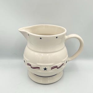 May include: Cream-colored ceramic pitcher with a rounded body and a handle. The pitcher is decorated with navy blue stars and red and navy blue ribbon-like designs. A classic piece for serving beverages.