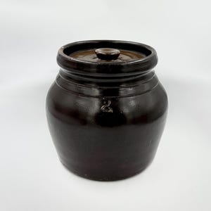 May include: A dark brown glazed stoneware crock with a matching lid and a small knob handle. The exterior of the cylindrical vessel features a prominent embossed number 2 on the front, indicating its capacity, with a smooth, glossy finish.