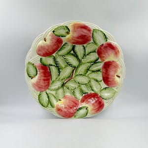 May include: A round ceramic plate featuring a raised, three-dimensional relief design of red apples and green leaves. The background of the plate has a cream-colored woven basket-weave texture, and the apples are arranged around the center.