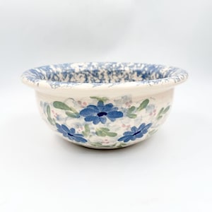 May include: A ceramic bowl with a cream-colored base featuring hand-painted blue flowers, green leaves, and small pink accents. The rim of the bowl is decorated with a blue and white speckled sponge-painted pattern around the entire circumference.