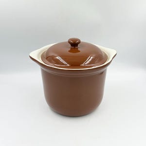 May include: A glossy brown ceramic pot with a matching lid and two small side handles. The pot features a smooth, rounded body and a simple knob on the center of the lid. The interior rim of the pot is visible, showing a contrasting cream color.
