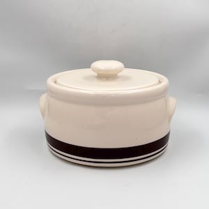 May include: Cream-colored ceramic casserole dish with a lid and two small handles. The dish has a dark brown band with two white stripes around the base. The lid has a small, round knob.