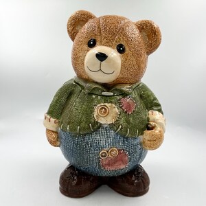 May include: A ceramic teddy bear cookie jar featuring a textured brown bear wearing a green sweater and blue pants with stitched patches. The bear has a cream-colored muzzle, black eyes, and a black nose. The sweater and pants have visible stitch details.