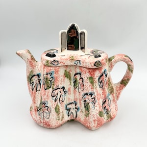 May include: A ceramic teapot with a fluted body, featuring a speckled pink and white base color. The surface is decorated with hand-painted black line drawings of flowers and green leaf accents. It has a matching lid with a central handle.