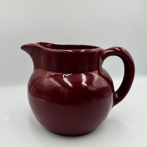 Könnte beinhalten: Ein bordeauxfarbener Keramikkrug mit rundem Körper, gebogenem Henkel und Ausguss. Der Krug hat eine glänzende Oberfläche und wird wahrscheinlich zum Servieren von Getränken verwendet. Das Design ist schlicht und klassisch.