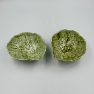 May include: Two small, glossy ceramic bowls shaped like green cabbage leaves. Each bowl features a detailed, embossed texture mimicking the veins and ruffled edges of a natural leaf, finished with a shiny, light to medium green glaze.