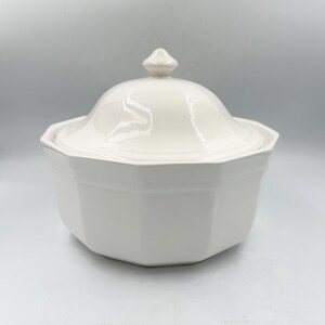 May include: A white ceramic sugar bowl with an octagonal base and a matching lid. The lid features a small, pointed finial at the center. The bowl has a smooth, glossy finish and a clean, geometric shape with eight distinct sides and edges.