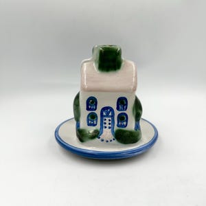 May include: A ceramic house-shaped incense burner or decorative figurine featuring a white exterior with a pale pink roof, a dark green chimney, and blue painted windows and door details, all resting on a matching circular white saucer base.