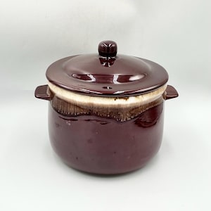 May include: A maroon ceramic casserole dish with a matching lid. The dish has a white and brown rim and two side handles. The lid has a small, rounded handle. The dish is likely vintage and suitable for cooking and serving.