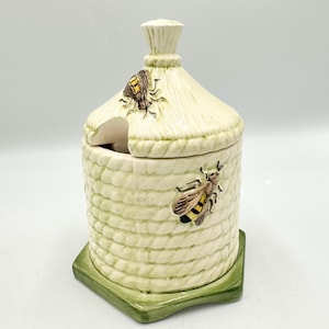 May include: A ceramic honey pot shaped like a beehive, with a straw-like roof and a green base. Two detailed bee figurines are attached to the pot. The pot is cream-colored with a rope-like texture.