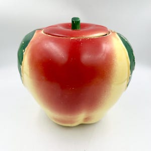 May include: A ceramic apple-shaped jar with a removable lid. The exterior features a gradient of red and cream colors with green leaf accents on the sides. A small green stem sits atop the center of the lid. The surface has a glossy finish.