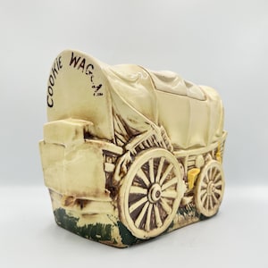 May include: Cream-colored ceramic cookie jar shaped like a covered wagon. The jar features brown wheels, a tan canvas top, and the words "COOKIE WAG-N" in brown lettering. The base has green and brown accents.