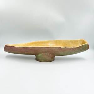 May include: An elongated ceramic bowl with a textured exterior featuring a blend of muted green and brown tones. The interior of the vessel has a smooth, glossy finish in a pale yellow hue. The bowl is supported by a central pedestal base.