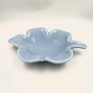 May include: A light blue ceramic dish shaped like a five-petaled flower. The bowl features a smooth, glossy finish with curved, raised edges that mimic the organic form of petals, creating a shallow basin for holding small items or trinkets.