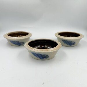 May include: Three round ceramic bowls with a cream-colored exterior and a dark brown interior. Each bowl features a scalloped rim and a hand-painted blue leaf-like design on the side. A small, centered protrusion is visible at the base of each bowl.
