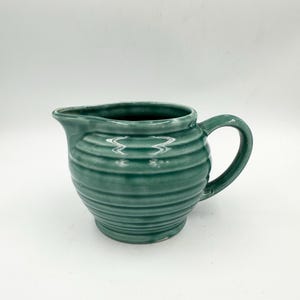 May include: A teal ceramic pitcher with a handle and a spout. The pitcher has a ribbed design around the body. The interior of the pitcher is a darker shade of teal. The pitcher is on a white background.