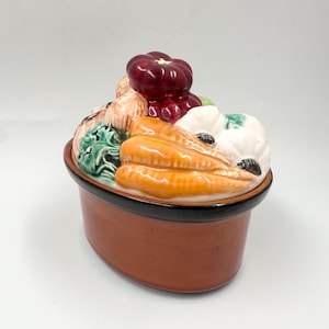 May include: A ceramic vegetable-shaped casserole dish with a brown base and black rim. The lid is adorned with colorful ceramic vegetables, including carrots, tomatoes, and garlic. The dish is designed to resemble a basket of fresh produce.