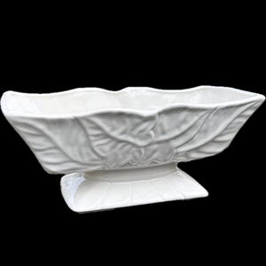 May include: A white ceramic serving dish with a leaf design. The dish is rectangular with a scalloped edge and sits on a square pedestal base. The dish is approximately 12 inches long.
