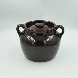 May include: A dark brown glazed ceramic bean pot with a matching lid and two side handles. The pot features a rounded body and a smooth, glossy finish. The lid has a small central knob for lifting, and the two handles are positioned on opposite sides.