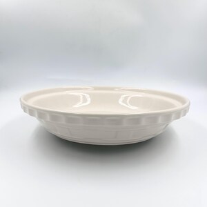 May include: A round, off-white ceramic pie dish with a smooth, glossy finish. The rim features a decorative, repeating fluted pattern that extends around the entire circumference of the dish, providing a textured edge to the simple, shallow bowl.