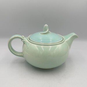 May include: A ceramic teapot with a rounded body and a light green glaze. The lid is a lighter shade of green and has a decorative handle. The teapot has a curved handle and a spout for pouring.