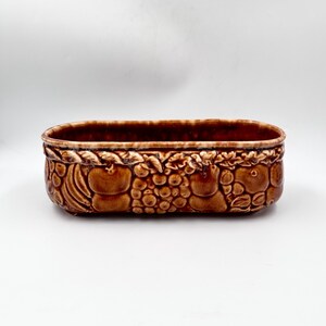 May include: An oblong ceramic planter with a glossy brown glaze. The exterior features a relief pattern of various fruits, including grapes and apples, encircling the entire side. The rim has a smooth, rounded edge with a slightly lighter brown tone.