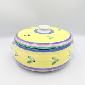 Puede incluir: Una fuente de cerámica amarilla con tapa. El plato tiene un asa blanca a cada lado y está decorado con formas de hojas verdes y rayas coloridas rosas, verdes y azules. La tapa tiene un pomo blanco.