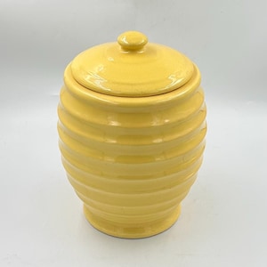 May include: A bright yellow ceramic honey pot with a beehive-inspired ribbed texture and a matching removable lid featuring a small knob handle. The container has a rounded body that tapers toward the base and a smooth, glossy finish throughout.