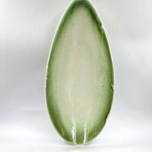 May include: An elongated, oval-shaped ceramic serving dish with a pointed tip. The interior features a smooth, off-white center that transitions into a textured, moss-green gradient along the curved edges, creating a distinct border effect.