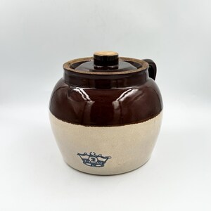 May include: A two-toned stoneware crock with a dark brown glazed upper section and a light beige lower section. It features a matching lid with a central knob, a side handle, and a blue stamped crown logo with the number 2 on the front body.
